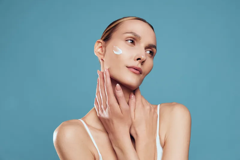 Young woman applies facial cream to her cheek, looking up with a calm expression, wearing a white tank top, isolated on a blue studio backdrop.