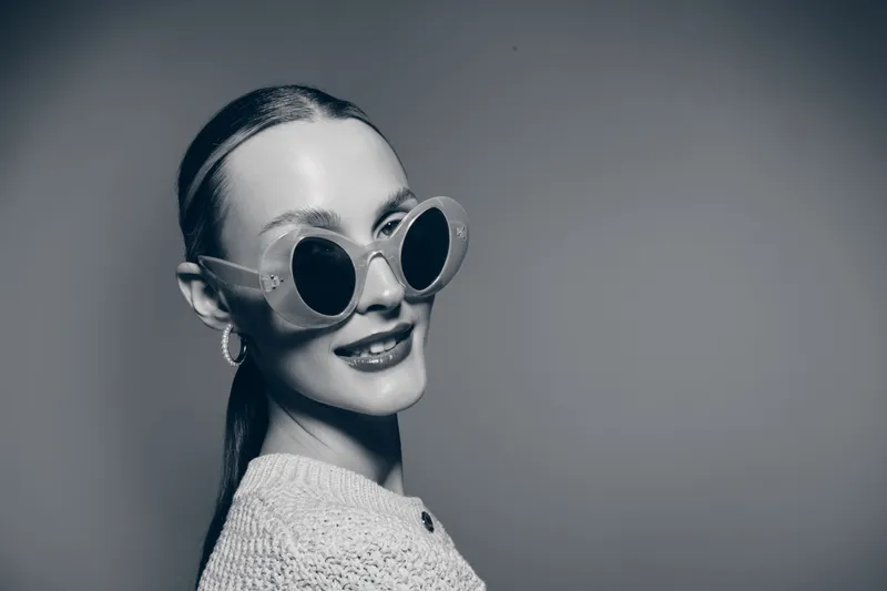 Young white woman with fair skin wearing round sunglasses and knitted sweater looks confident and stylish on plain background in black and white portrait.