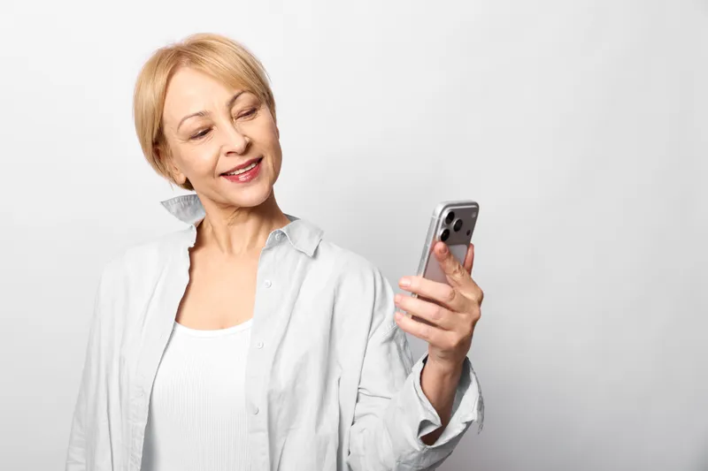 Smiling mature woman indoors against a plain white background, holding a smartphone and looking at the screen, relaxed posture, soft light, technology and communication moment