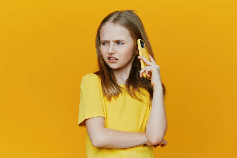 A young girl with straight blonde hair in a yellow t shirt, expressing confusion while talking on a phone against a vibrant yellow background