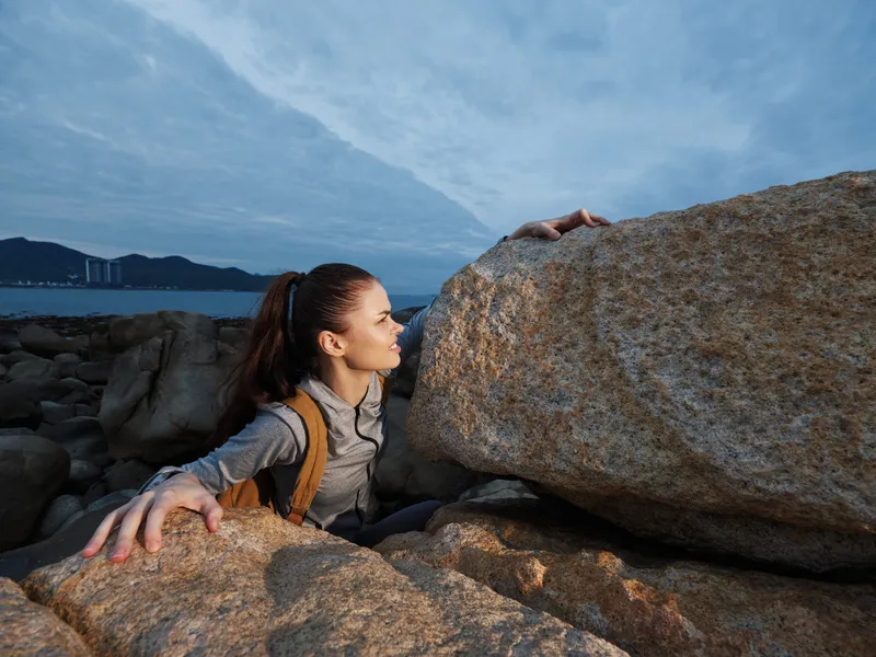 A young woman in athletic clothing climbing rocky terrain by the sea, showcasing determination and strength against a cloudy sky at dawn, ideal for fitness or adventure themes