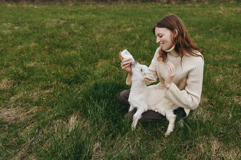 Woman outdoors feeds a young goat kid with a bottle in a verdant pasture, a tender bonding moment that highlights nurturing care, calm joy, and simple rustic life under bright natural light.