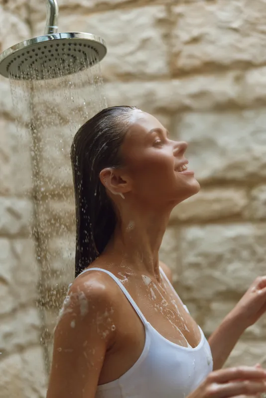 Woman enjoying outdoor shower with closed eyes, water flowing on face and shoulders, natural stone background, summer wellness moment, peaceful mood