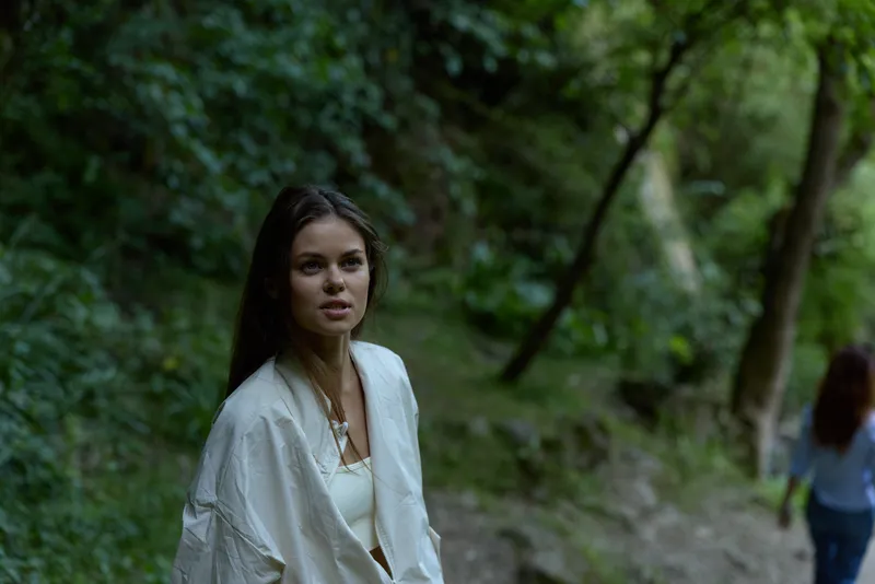 young woman in white jacket standing in lush green forest with thoughtful expression and soft natural light, peaceful outdoor scene