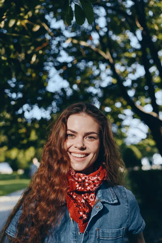 Smiling woman in modern boho-western denim street style with curly hair and red bandana, captured in warm film color lighting outdoors under lush green trees.