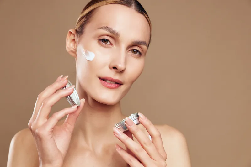 Young woman applying moisturizing cream on face, gentle skincare routine, soft natural light, smooth skin texture, neutral background, beauty and health concept