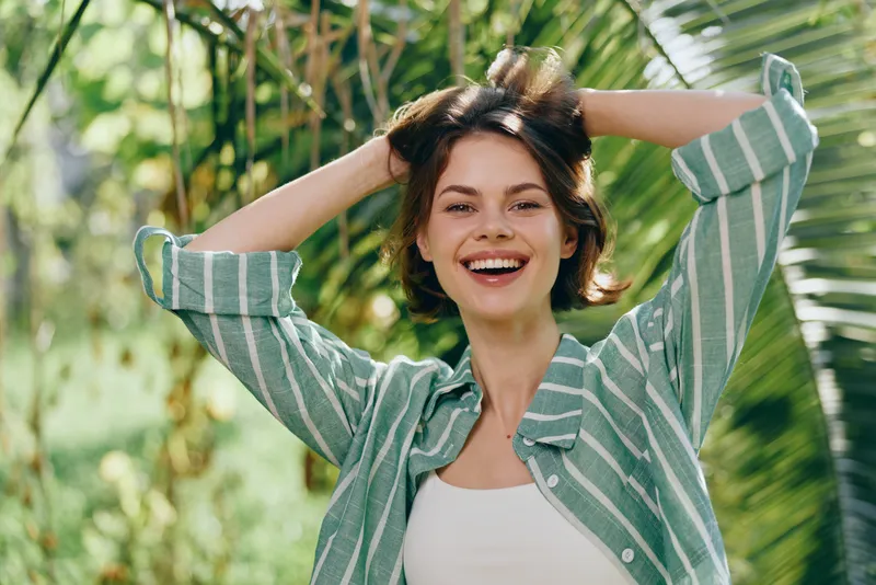 woman smiling outdoors in bright daylight, a cheerful woman wearing a striped green shirt raises her arms, capturing a sunny, joyful moment in nature.
