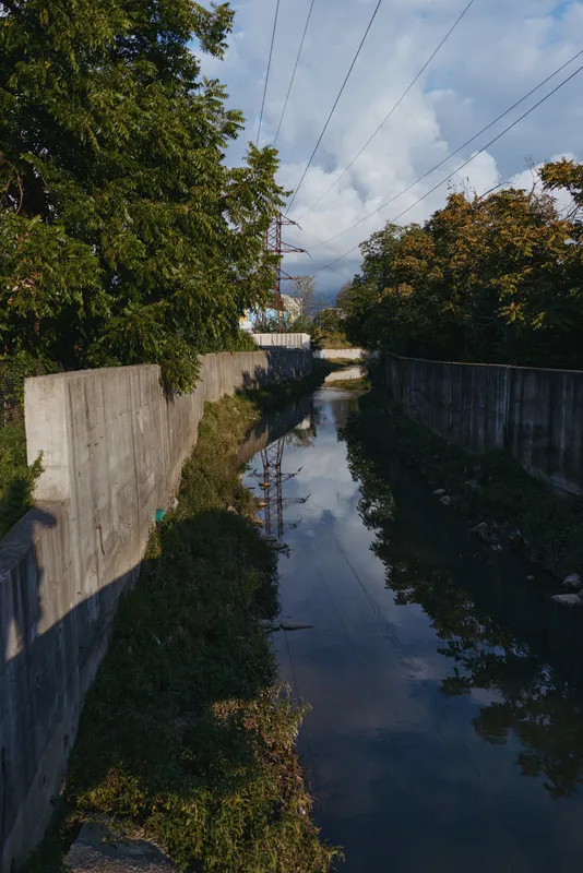 Concrete canal, drainage channel, reflection, trees, power lines, shadow — a narrow suburban drainage with an adult male shadow walking in late afternoon light, cloudy sky and moody travel atmosphere.