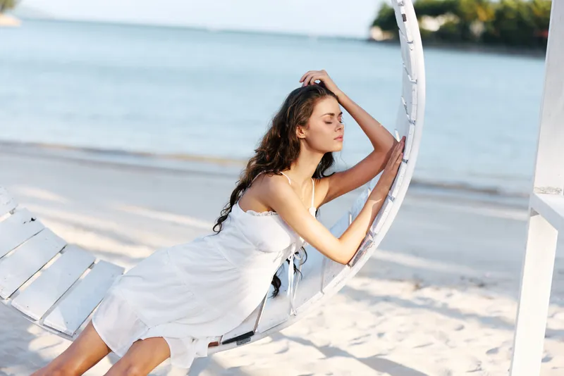 A young woman in a white dress leans on a circular seaside swing, gazing toward the horizon as soft waves roll onto the sandy shore.