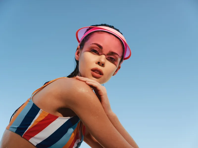 Portrait of a young woman in a striped swimsuit and pink visor, sunlit skin and confident expression, captured from a low angle against a clear blue sky, emphasizing summer vitality and effortless
