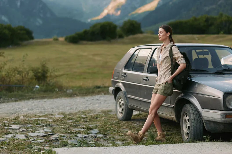 A woman on a road trip rests against a rugged car in a scenic countryside scene, exuding casual confidence and wanderlust as mountains rise in the background.