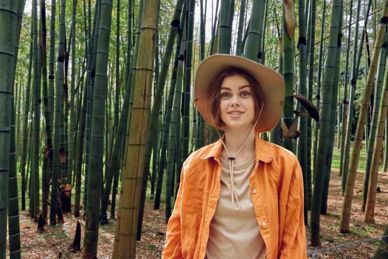 woman hat bamboo forest portrait travel: Smiling woman in an orange jacket and wide brim hat stands among tall bamboo stalks in a serene forest, a casual travel portrait with warm natural sunlight