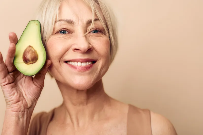 Smiling mature woman with short blonde hair holds half avocado near face on beige background. Concept of healthy lifestyle, natural skincare, nutrition, and wellness for seniors.