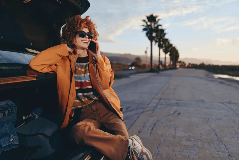 Woman lifestyle, curly hair, orange jacket, sunglasses, sitting by car on road, smiling and talking on phone, travel at sunset, warm outdoor light, casual travel mood