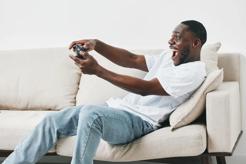 Happy young African American man playing video games on a couch, showcasing excitement and engagement with the gaming experience