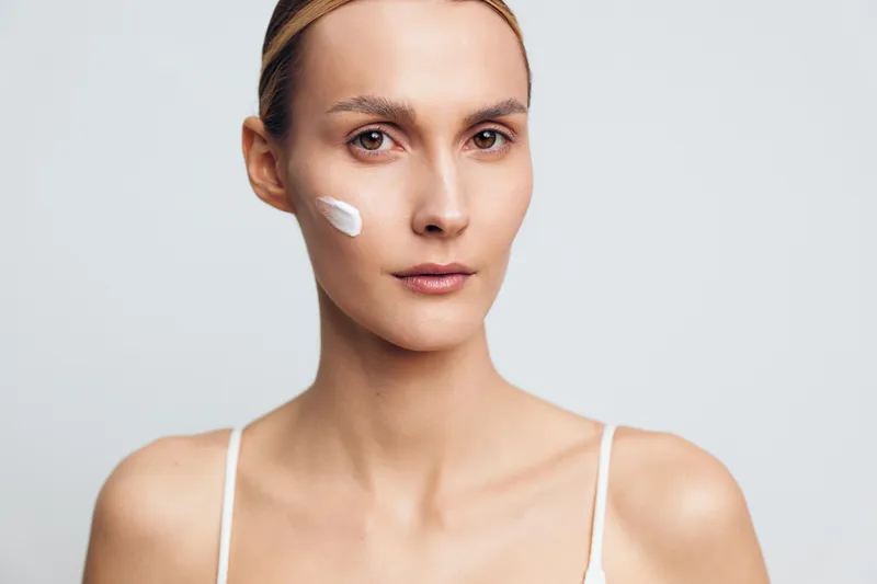 Young woman with clean skin applies white facial cream on cheek looking at camera with calm expression isolated on light background in skincare beauty concept.