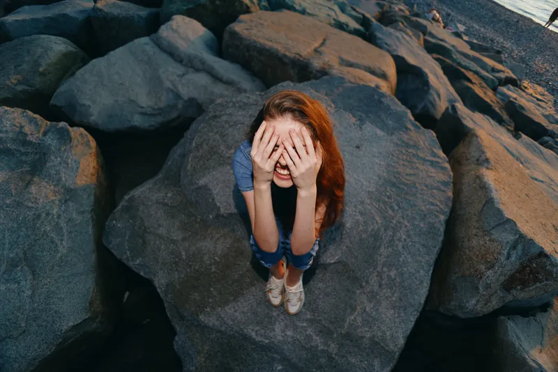 redhead woman covering face sitting on large rocks by the water, casual clothes, outdoor, natural light, candid moment, emotional expression