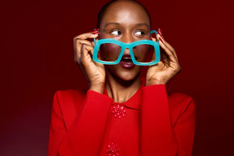 Portrait of a young black woman with dark skin wearing a red sweater holding large turquoise glasses near her face on a red background. Fashion concept with vibrant colors and style.