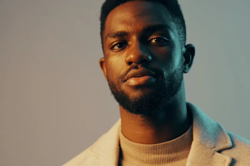 A confident young Black man in a stylish gray coat and beige turtleneck, portraying a sense of sophistication against a cool, gradient background