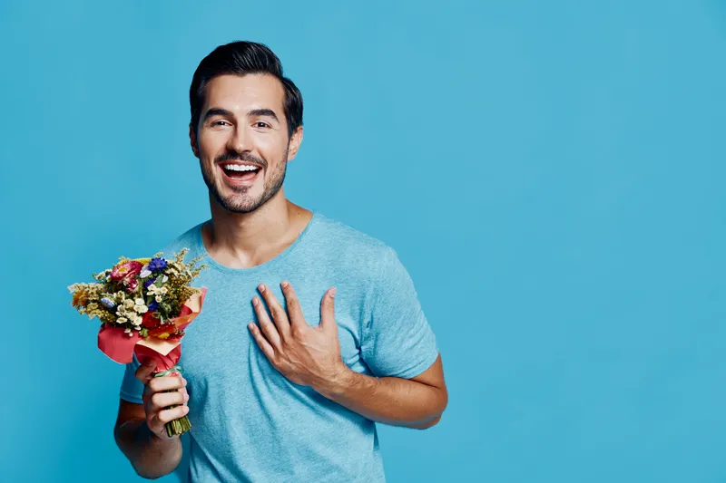 Happy smiling man holding a bouquet of flowers, wearing a casual blue t-shirt, standing against a solid turquoise background, expressing joy and celebration.