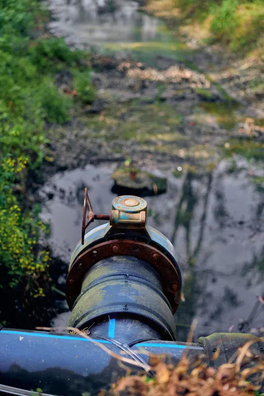 Pipe valve drain releasing water into a muddy stream, pollution and rust visible on metal fittings, wastewater discharge in wetland with vegetation and contaminated runoff and puddles.