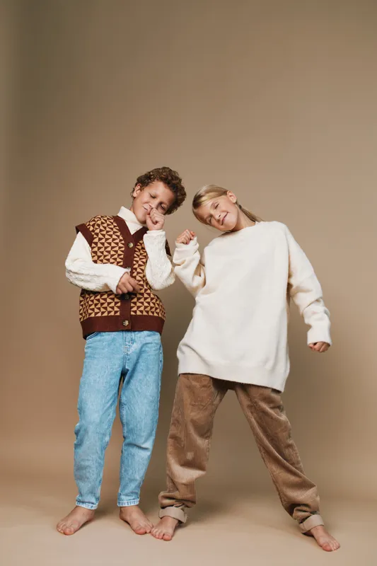 Two friends wear cozy knitwear and relax on a soft beige backdrop, posing playfully with smiles, showcasing casual comfort, warmth, and relaxed fashion style.
