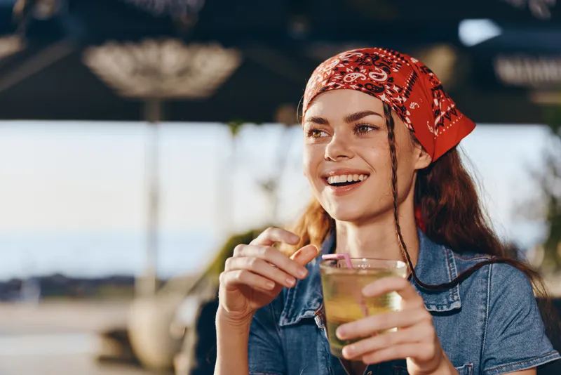 smiling woman with red bandana holding a cocktail glass enjoying a sunny outdoor setting with casual denim shirt and blurred background