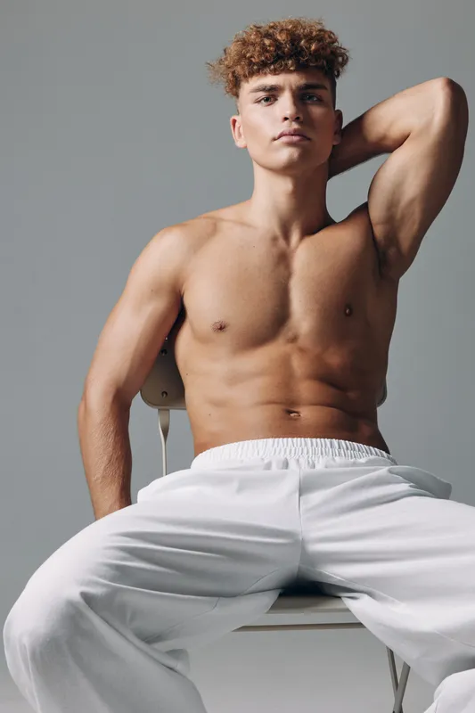 Young athletic man with muscular torso posing shirtless sitting on chair against plain background for fitness, wellness and healthy lifestyle concept in studio.