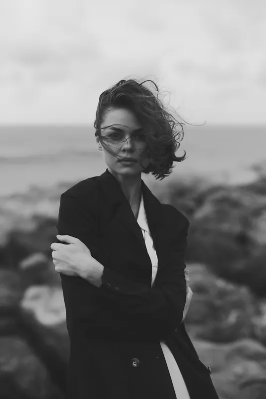 Monochrome fashion shot of a windblown woman in a tailored coat by the sea, sunglasses on, embodying chic style and bold attitude on a rugged beach.