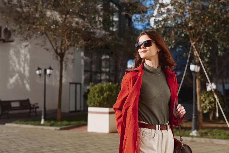 A confident young woman wearing sunglasses and a red trench coat walks outdoors on a sunny autumn day with trees and street lamps in the background.