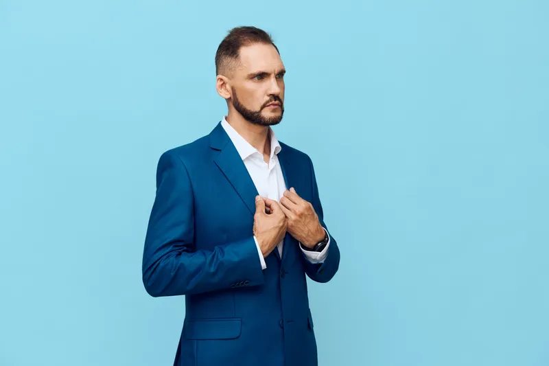 A professional man in a blue suit stands against a solid blue background, exuding confidence and focus as he adjusts his jacket, signaling readiness and calm determination.
