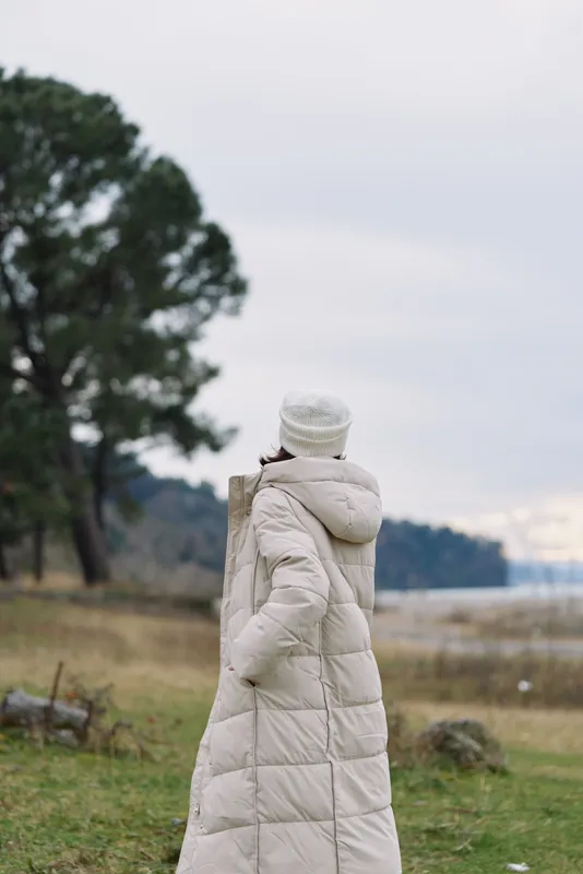Woman in a long beige puffer coat stands in an open field beside a lake, with trees on the horizon, conveying a serene winter outdoor scene and quiet breeze.