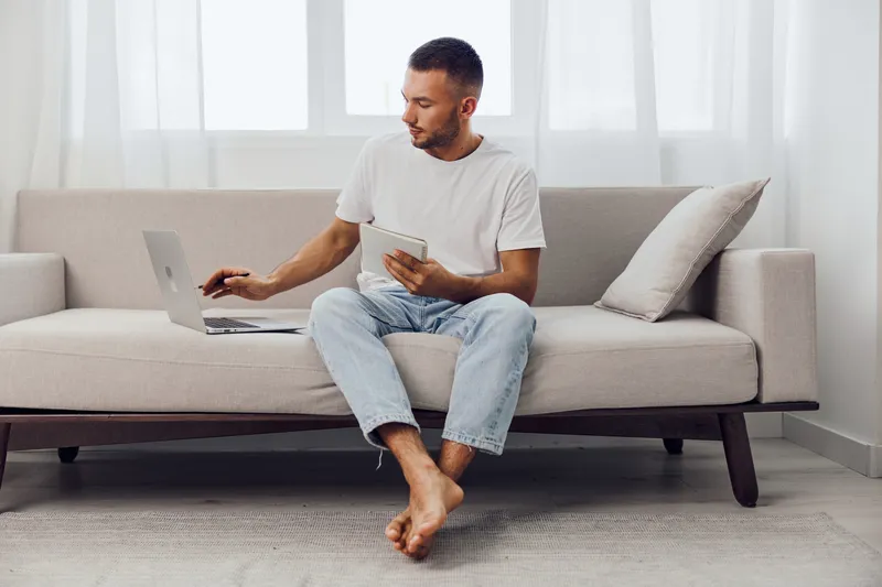 A focused man working on a laptop while holding a tablet, showcasing a modern home office setup. Bright, airy atmosphere enhances productivity and creativity.