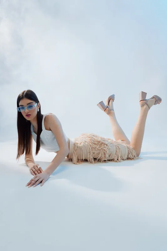 Young woman in a white top and fringed skirt poses lying down on a light background wearing sunglasses and high heels. Concept of beauty fashion and modern style.