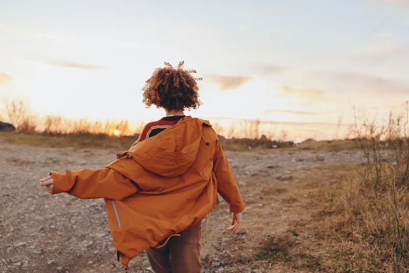 Child boy in orange jacket with hood walking outdoors at sunset, lifestyle moment in open field, rear view, warm evening light, soft clouds, fresh air and calm mood for childhood exploration