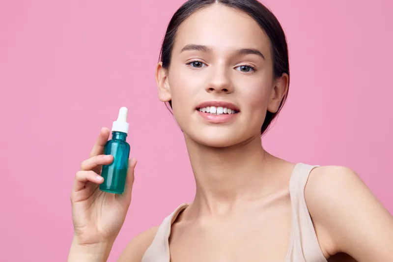 A young woman holding a green dropper bottle in a beige top, smiling against a pink background, showcasing skincare and healthy beauty concepts