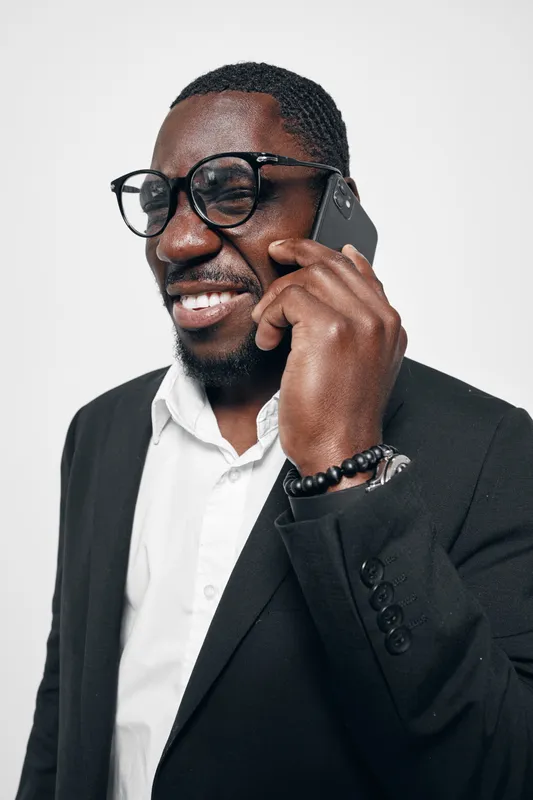 African American man with dark skin wearing glasses and black suit talking on smartphone smiling against white background.
