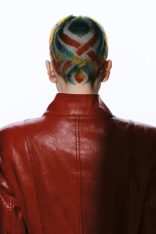 Rear view of a woman with colorful artistic shaved hair design wearing a shiny red leather jacket, showcasing unique style and bold fashion choice.