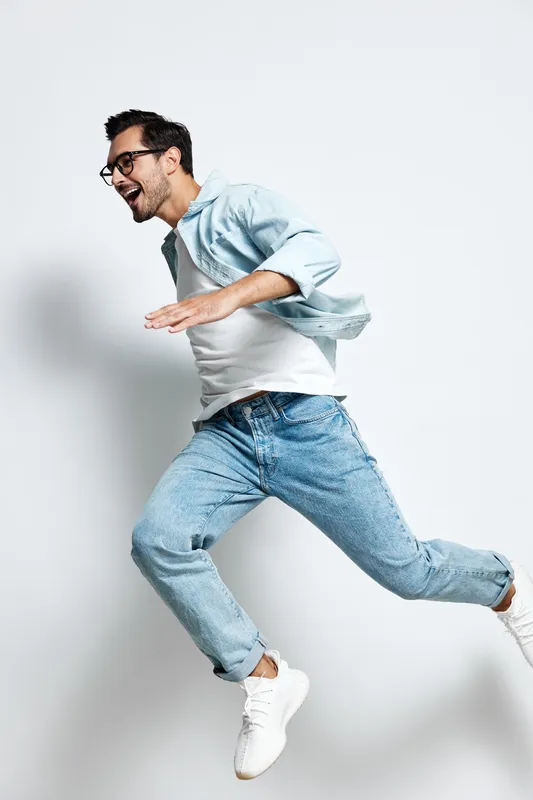Full body joyful happy young man jumping in light denim outfit with glasses, expressing excitement and energy. Isolated on plain light background studio portrait.