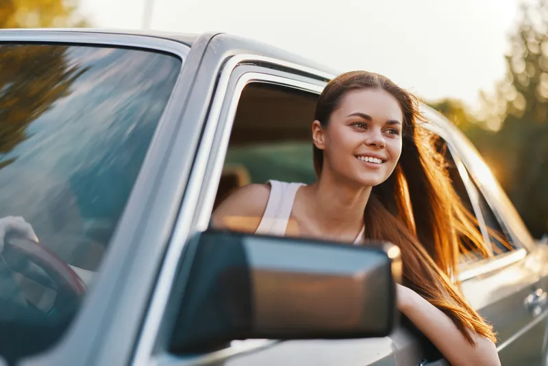 Young woman enjoying a road trip with her hair blowing in the wind, smiling out of a car window on a sunny day, conveying freedom and happiness.