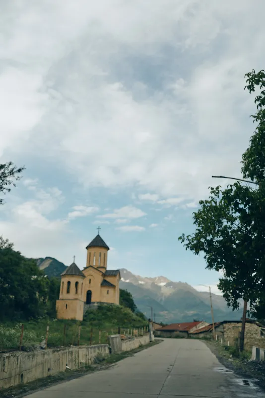 Countryside road leads to a small church set against towering mountains under a bright sky, creating a serene landscape with rural charm and peaceful village atmosphere