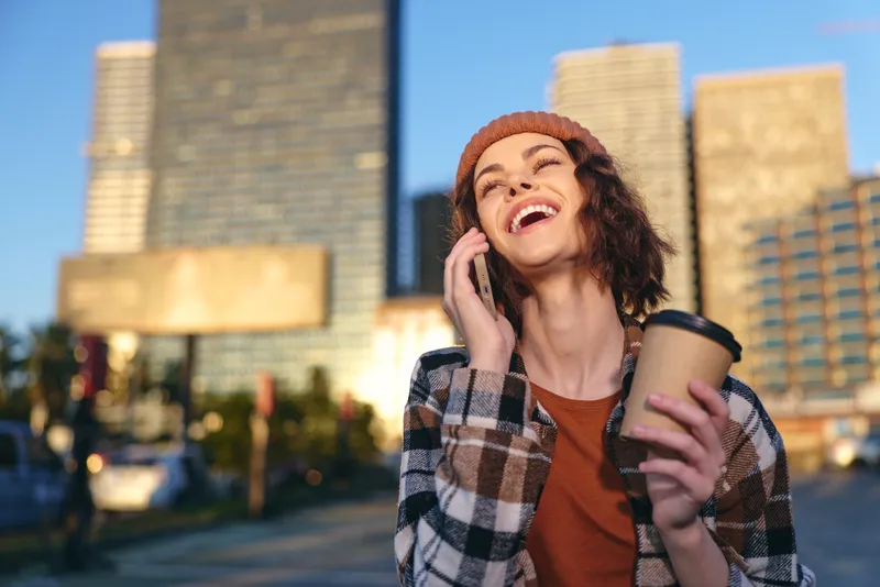 woman city coffee phone smiling urban candid lifestyle authenticity captured in golden hour glow, mindful living and emotional storytelling as she laughs holding a takeaway cup outdoors.