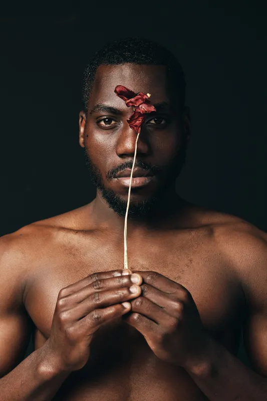 Creative conceptual portrait of African man holding dried flower in front of face with dark background, artistic visual style, closeup, bare chest, thoughtful expression.