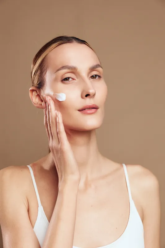 Young woman with clear skin applying white face cream on cheek. Close-up portrait with neutral background. Concept of skincare, beauty routine, self-care, and natural wellness.