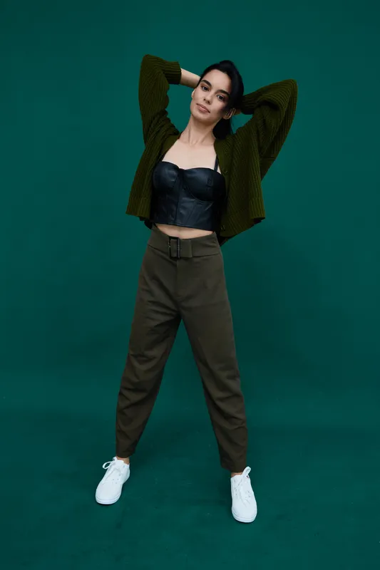 Young woman posing confidently in casual autumn outfit with green cardigan, black leather top, olive pants and white sneakers against dark green background.