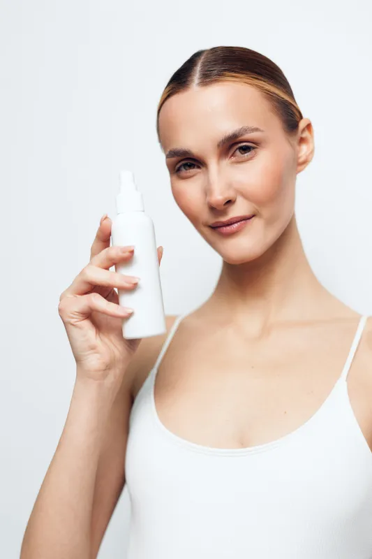 Young woman with clear skin holding a white cosmetic spray bottle, wearing a white tank top, standing against a plain light background, concept of skincare and beauty routine.