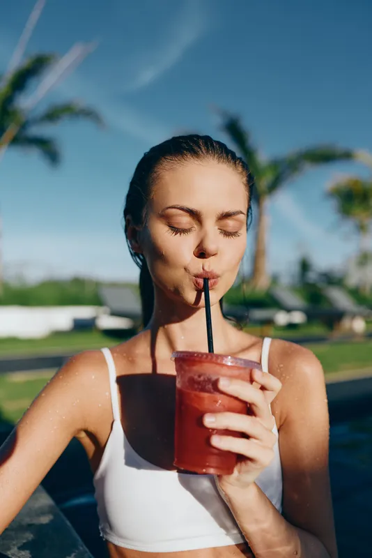Cheerful woman by a pool enjoying a refreshing smoothie, sunlit moment, relaxed pose, tropical backdrop, blue sky, palm trees, vibrant colors, summer mood