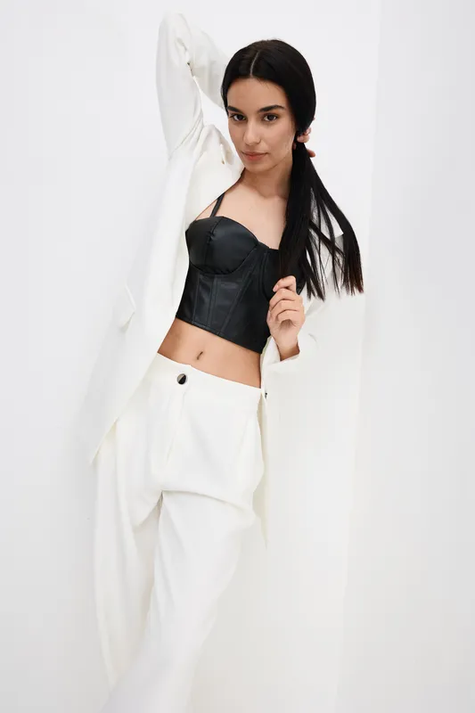 Young woman with black corset and white pants posing confidently against a white background, showcasing modern fashion and sleek hairstyle in a minimalist studio setting.