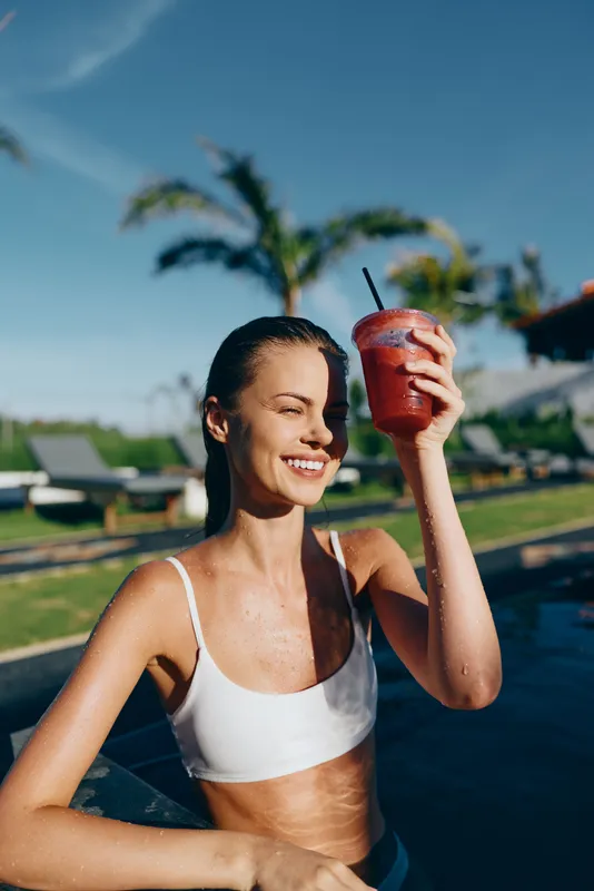 Young woman in a white bikini top enjoys a refreshing red smoothie by the pool on a sunny day, smiling happily and shading her eyes with the drink.