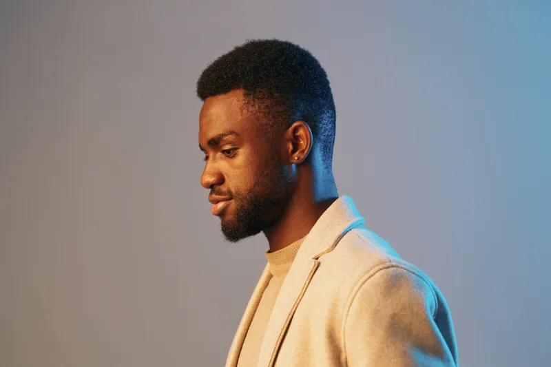 Profile of a stylish Black man in a light gray blazer, showcasing thoughtful expression against a gradient blue background, ideal for concepts of fashion and modern lifestyle